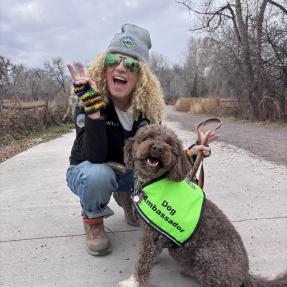 a woman wearing an OSMP beanie hat poses for a picture on a trail with her brown dog wearing a neon yellow vest that says "Dog Ambassador".