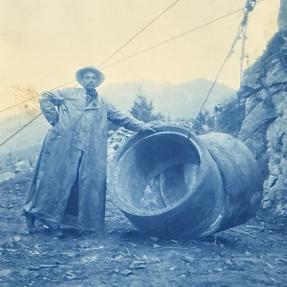 Undated, a man standing near sections of pipe being hoisted by the tram system. 