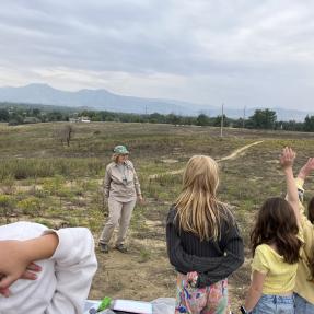 Standing in an open field, a volunteer in uniform faces a group of students with hands raised.