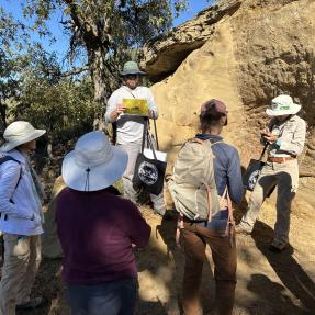 Two people presenting with photos to a group next to a smooth cliff.