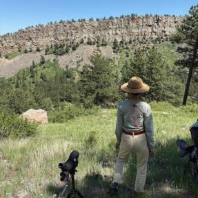 A person stands on a grassy hillside facing a rock cliff with a spotting scope.