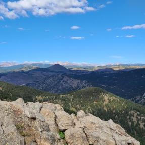A viewscape of an area as seen from a Boulder OSMP trail. In the foreground there are large, bare rocks. The middle distance is made up of mountains covered in green conifers, adn the far distance depicts snow-topped mountains.  