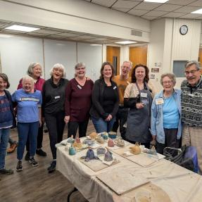Group of older adults posing behind a table displaying their finished pottery pieces. 