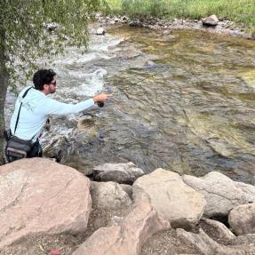 Man with arm stretched out to cast a fishing line over a full river with trees and rocks surrounding the river.