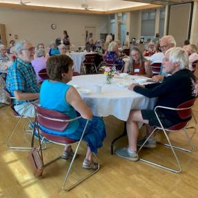 Groups of older adults sitting around tables conversing, in the East Age Well Center. 