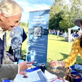 A man reviews materials at the Community Mediation and Resolution Center information table while a smiling woman in a yellow shirt assists him at a sunny outdoor information fair.