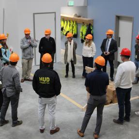 Approximately 15 tour participants wearing orange and red hard hats gather in a loose circle inside BoulderMOD for a group discussion. 