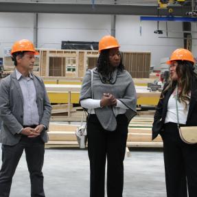 Three visitors wearing orange hard hats converse on the floor of the BoulderMOD, with partially framed modular housing panels and construction equipment visible in the background.