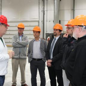 A person in a red hard hat and white jacket leads a discussion with five visitors wearing orange hard hats inside BoulderMOD.