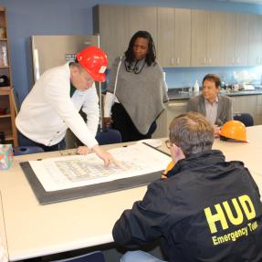 Dan McColley, Executive Director of Flatirons Habitat for Humanity, leans over a conference table to point out details on a large printed site map, while several others look on in a break room setting.