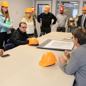 A group of visitors wearing orange hard hats gathers around a conference table during a briefing at BoulderMOD.