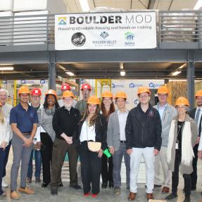 A group of approximately 15 adults wearing orange hard hats pose for a group photo inside BoulderMOD. Behind them hangs a banner reading "BoulderMOD – Providing affordable housing and hands on training," featuring logos for the City of Boulder, Boulder Valley School District, and Flatirons Habitat for Humanity. 