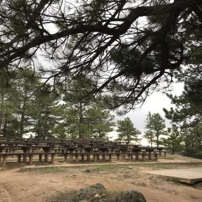 Several rows of picnic tables spread out from a small stage platform with pine trees in the background.