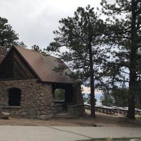 The Stone Shelter surrounded by pine trees with picnic tables in the background.