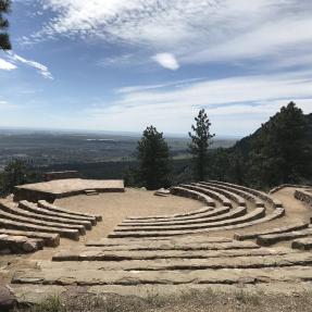 Rows of stone seats arc around the center circle and stage of the amphitheater, with the Flatirons visible to the right.