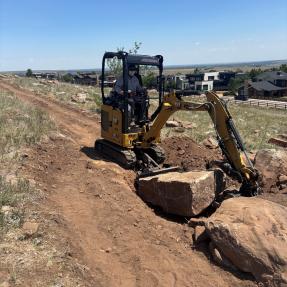 A person sits in a small construction equipment vehicle moving a large rock.