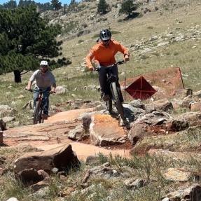 Two mountain bikers riding over a large rocks on the North Foothills Trail
