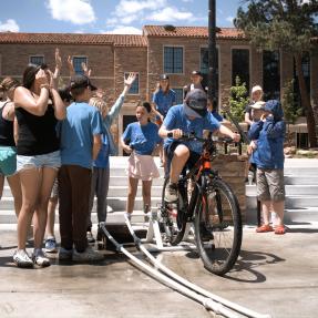 kids in blue shirts with hands outstretched to catch water as one kid pedals on a bike