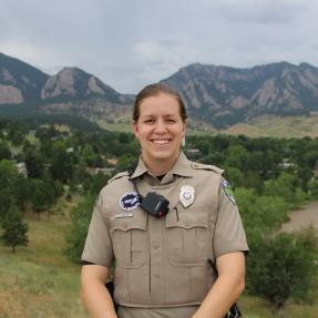Urban Ranger Caitlyn Imfeld stands above Harlow Platts Park, with the Boulder Flatirons and Viele Lake visible in the background. 