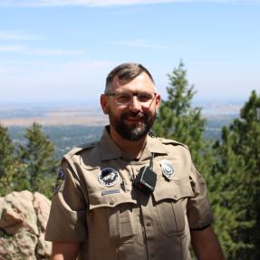 Urban Ranger Brian Bauer stands on top of Flagstaff Mountain, framed by trees and blue skies, with a view of Boulder in the background. 