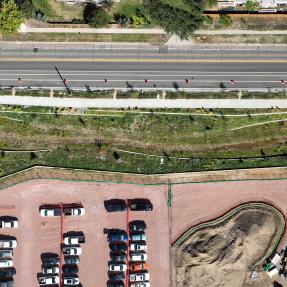 Aerial view straight down on the long, terraced flood channel running between the roadway and a construction site with a gravel parking lot.