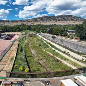 Grassy Alpine-Balsam flood channel with concrete walls, seen from a bridge railing, with a sidewalk and road on the right, a gravel parking area on the left, and foothills beyond.