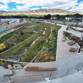 Curving flood channel with native grasses, wildflowers, and a thin stream of water, beside a brick-paver plaza with wooden benches, boulders, and flowering plantings.