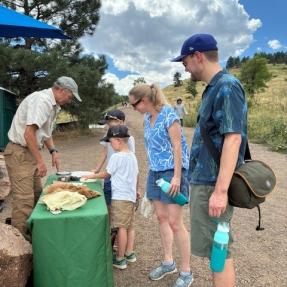 A family with young kids looking at pelts on a table while a person stands on the other side of the table pointing at the pelt.