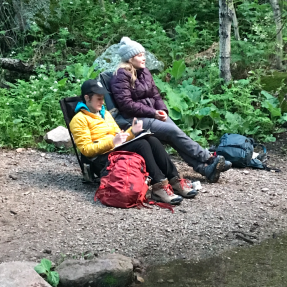 Two people sitting outside next to a body of water writing on a clipboard.