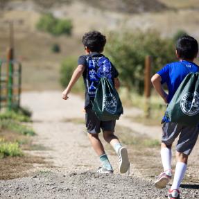 Two young kids with backpacks on run along a gravel trail outside.
