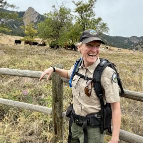 A woman in a tan uniform and backpack stands next to a field of cattle smiling.