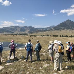 Several people standing in a rocky prairie looking out at a small pond.