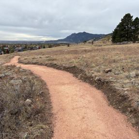 A smooth dirt trail winds downhill with a few of the Flatirons in the distance. 