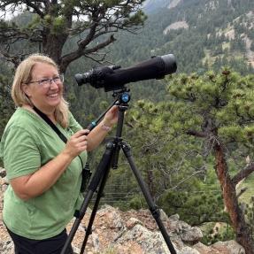 A woman standing on a mountain side with a very large lens camera on a tripod.