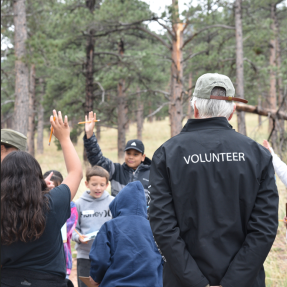 The back of a person with a jacket that reads 'Volunteer'. He is looking a bunch of kids with their hands raised.